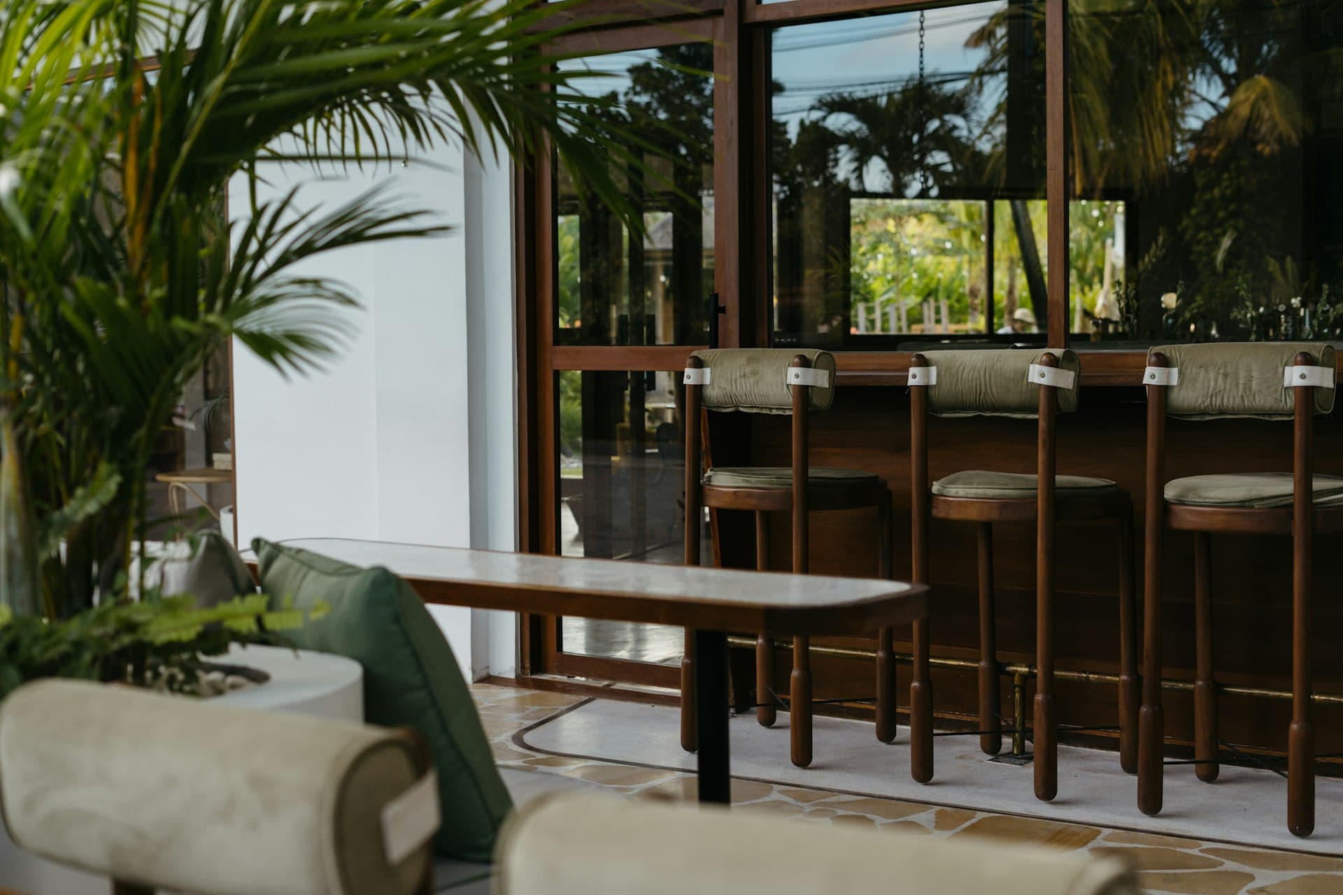 Modern cafe interior with stools at the counter, tropical plants, and large glass windows looking out to greenery