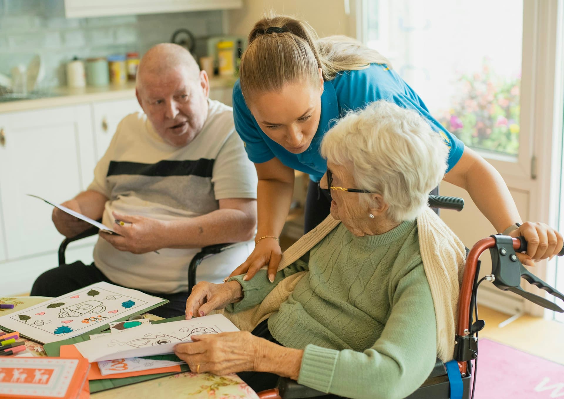 Home-health caregiver in scrubs assisting two elderly patients with an activity at a table in a residential care setting