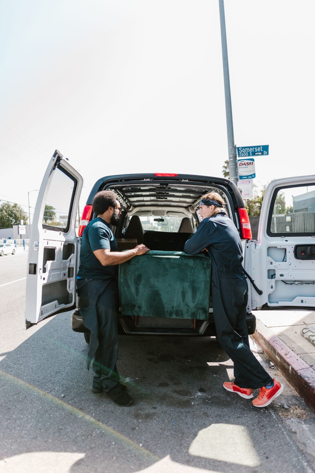 Two movers in plain work coveralls lifting a padded upholstered piece into the rear of a white cargo van on a city street