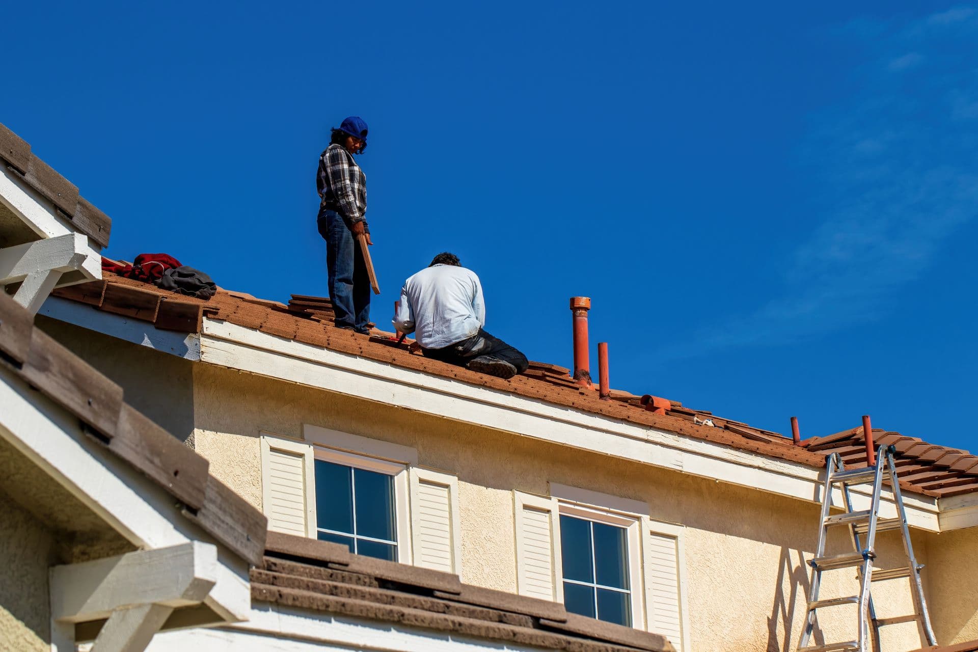Two roofers working on a residential tile roof under a clear sky, one carrying material while the other sets tiles