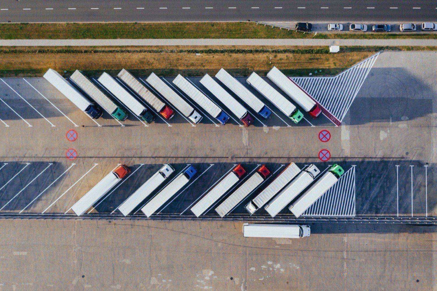 Aerial view of a fleet of semi-truck tractors with white trailers parked in diagonal rows at a commercial trucking yard