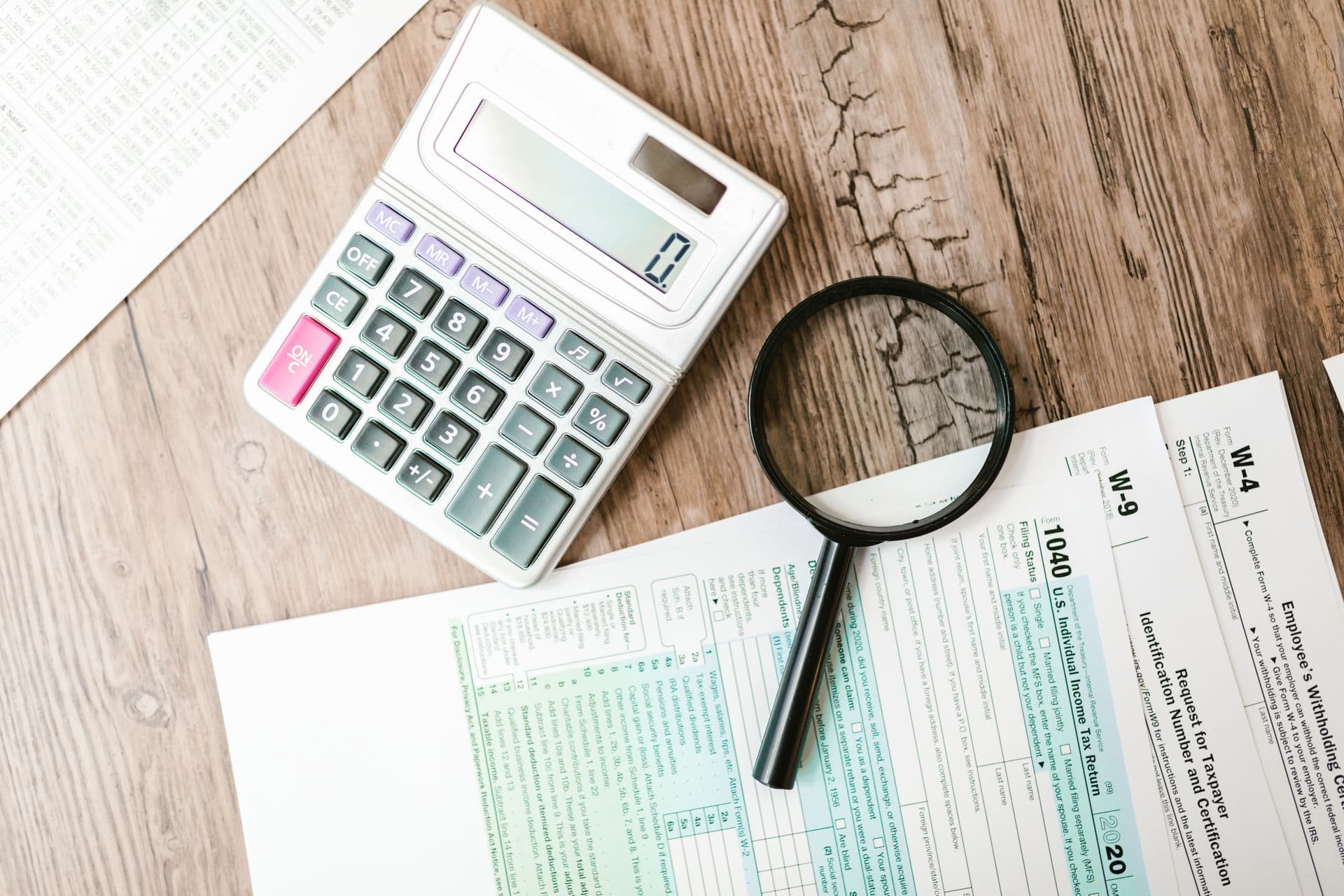 A calculator and magnifying glass laid over financial forms on a wooden desk, an underwriting review and audit workspace