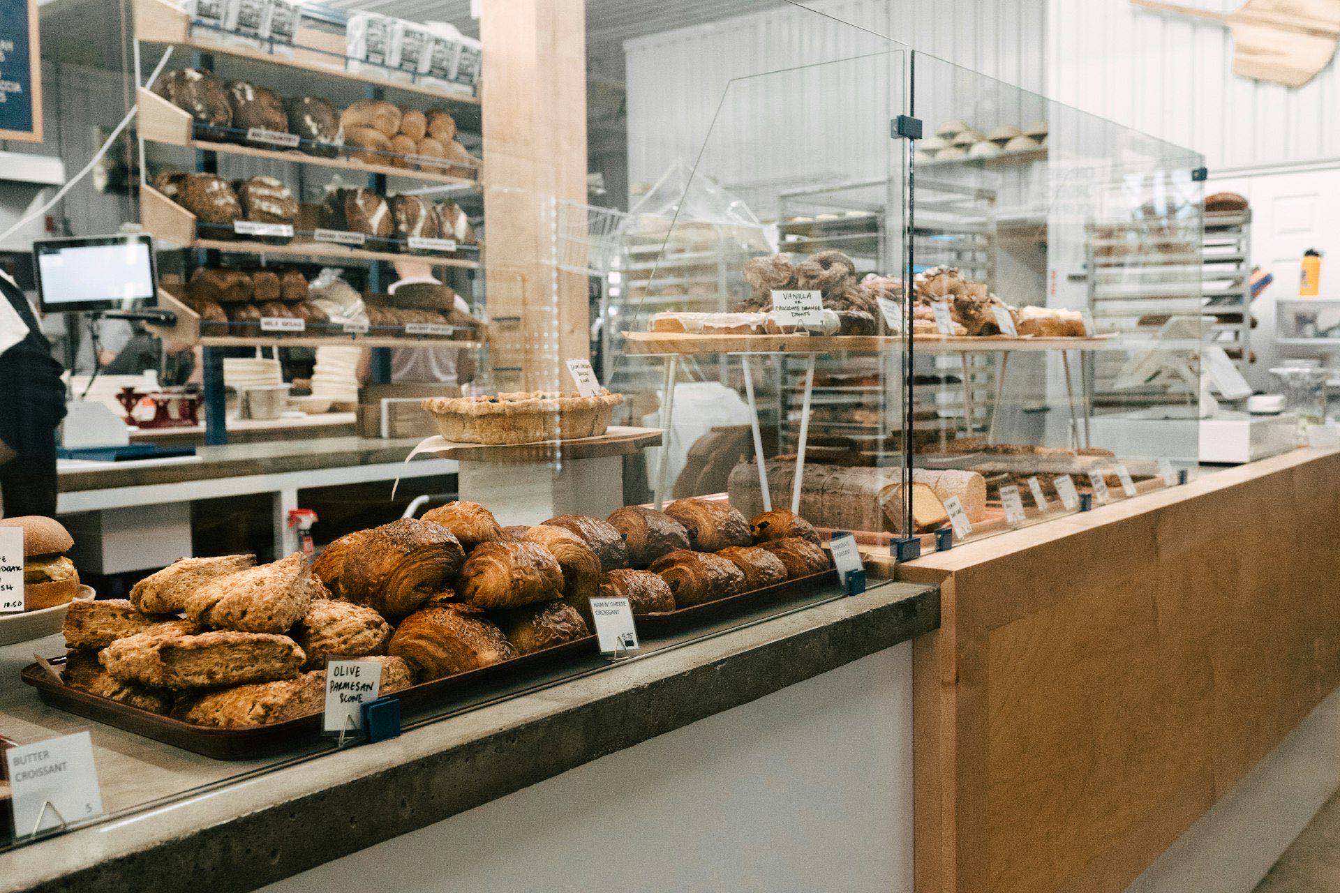 Artisan bakery counter with croissants, scones, and sourdough loaves arranged in trays, kitchen visible behind glass