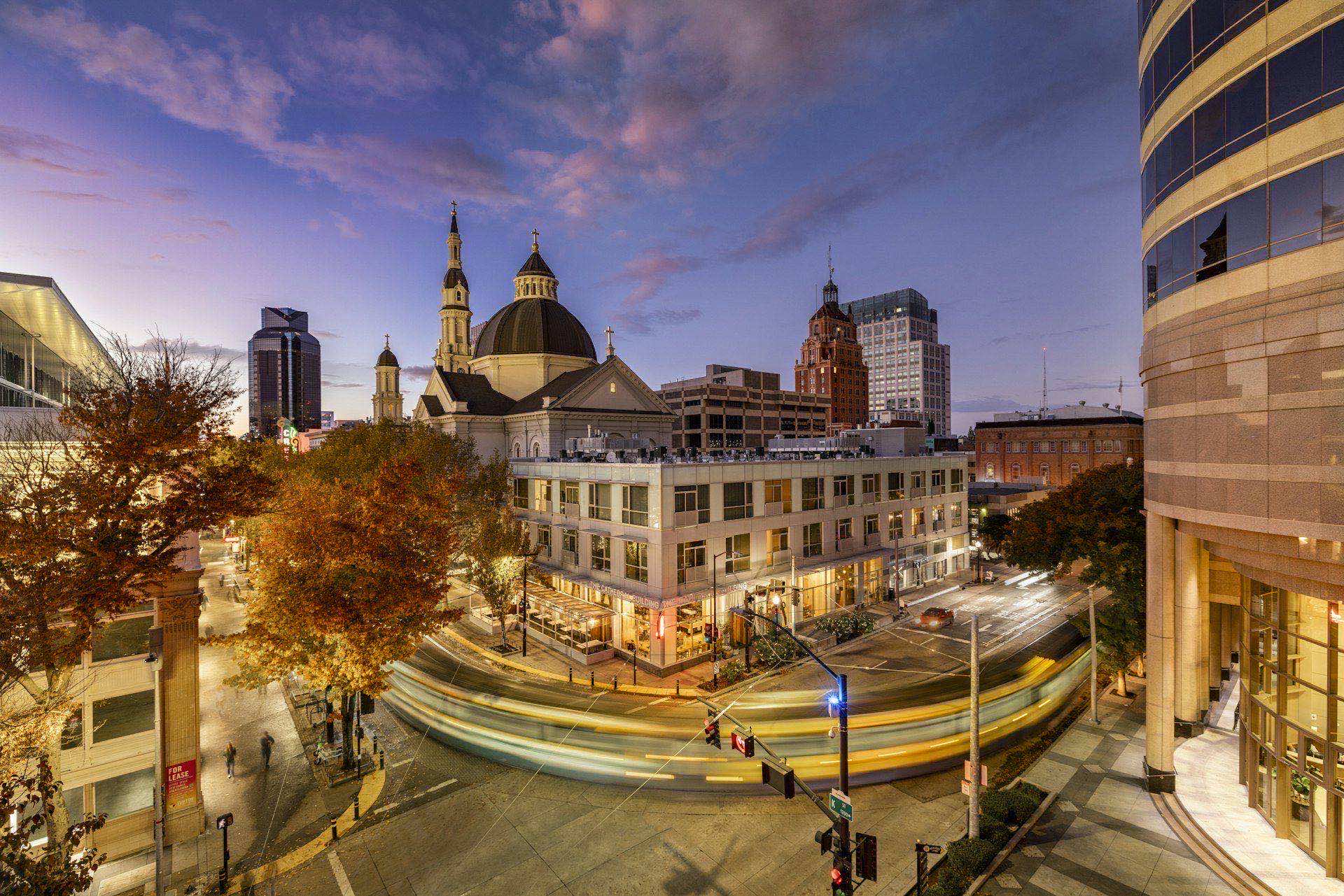 A downtown commercial district at dusk with light-trailed traffic curving past illuminated storefronts and towers