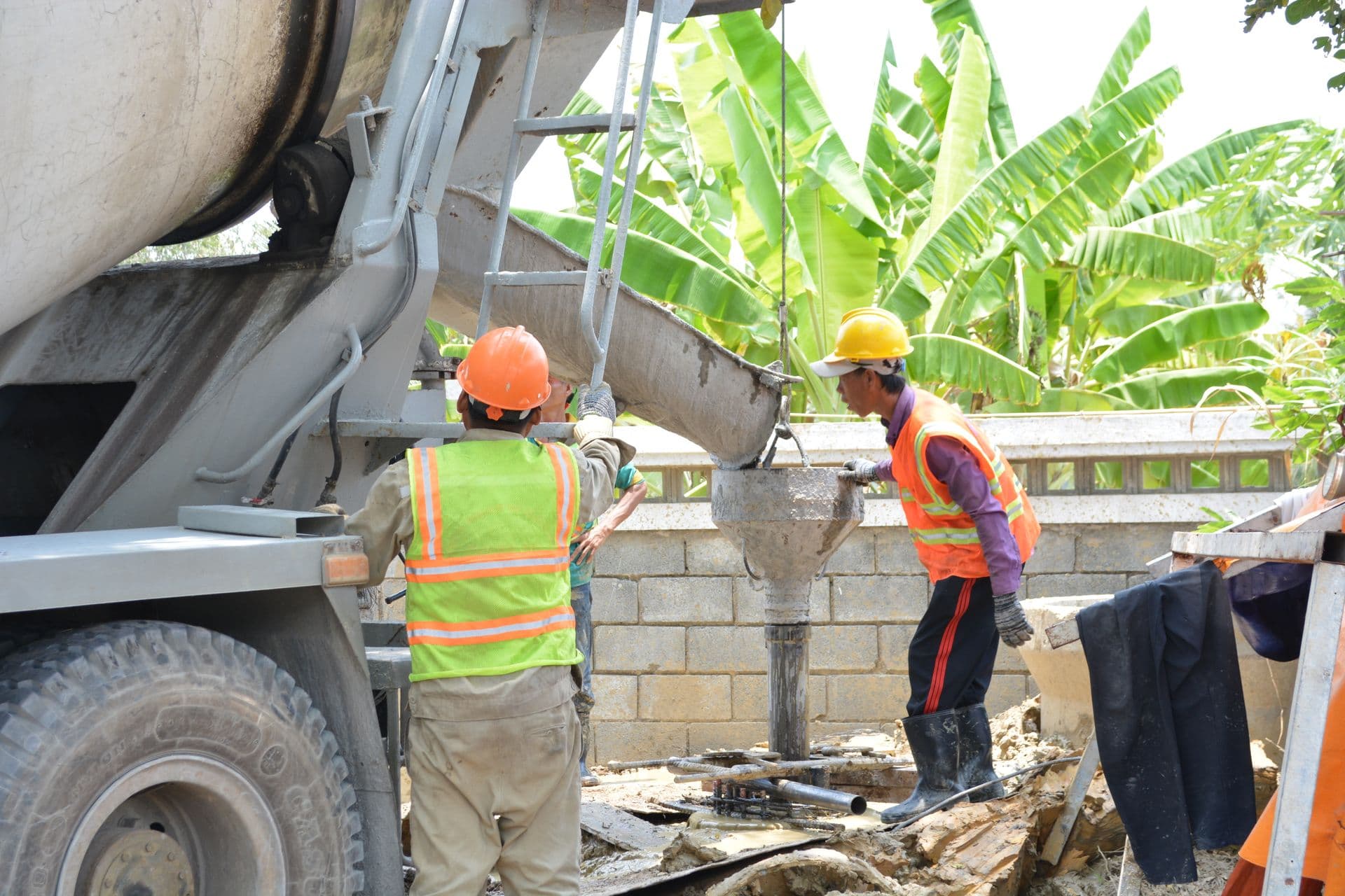 Two workers in hard hats and vests guiding concrete from a mixer-truck chute into a foundation form at an outdoor site