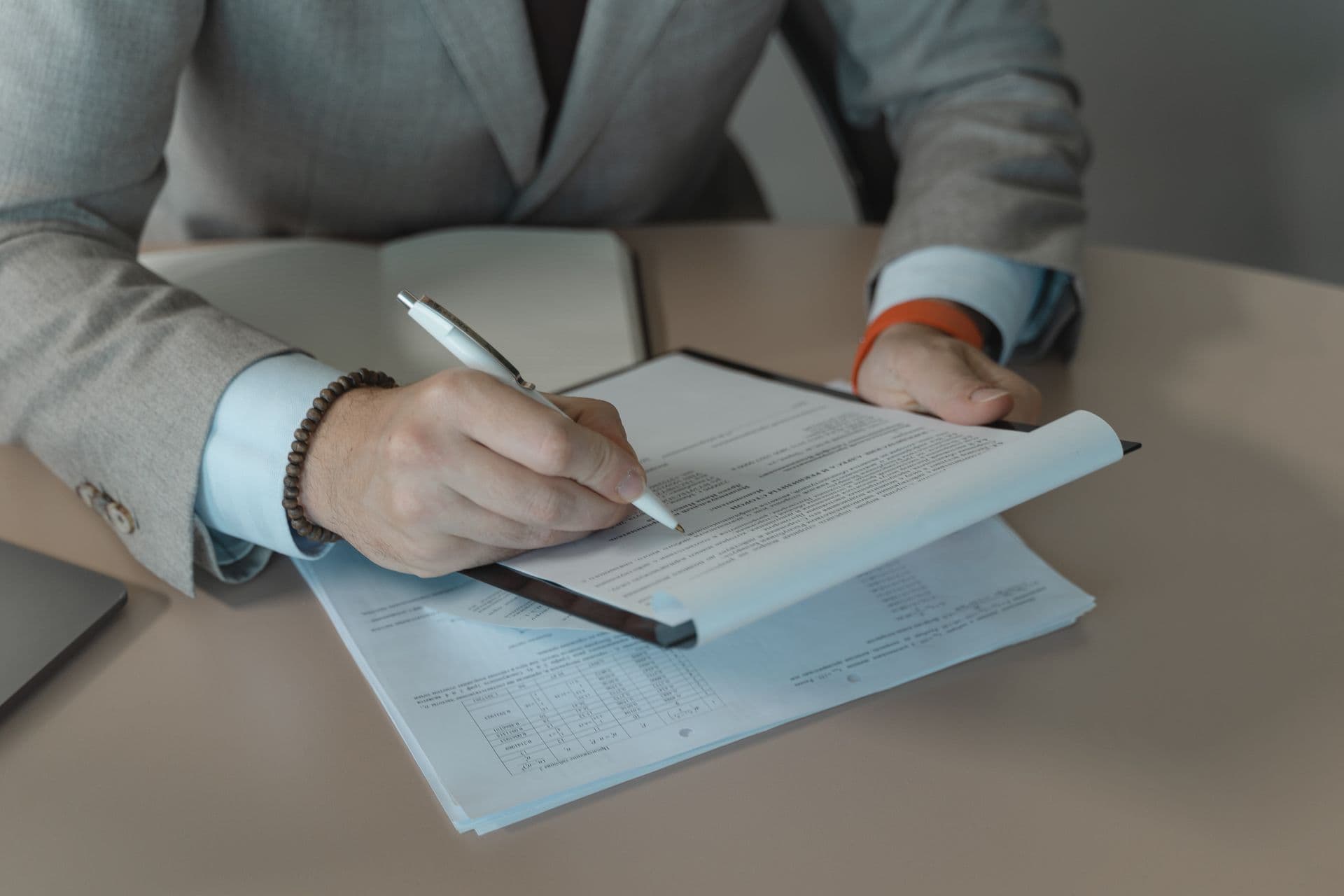 A person signs a multi-page contract clipped to a dark clipboard on a desk, pen in hand — certificate and contract paperwork