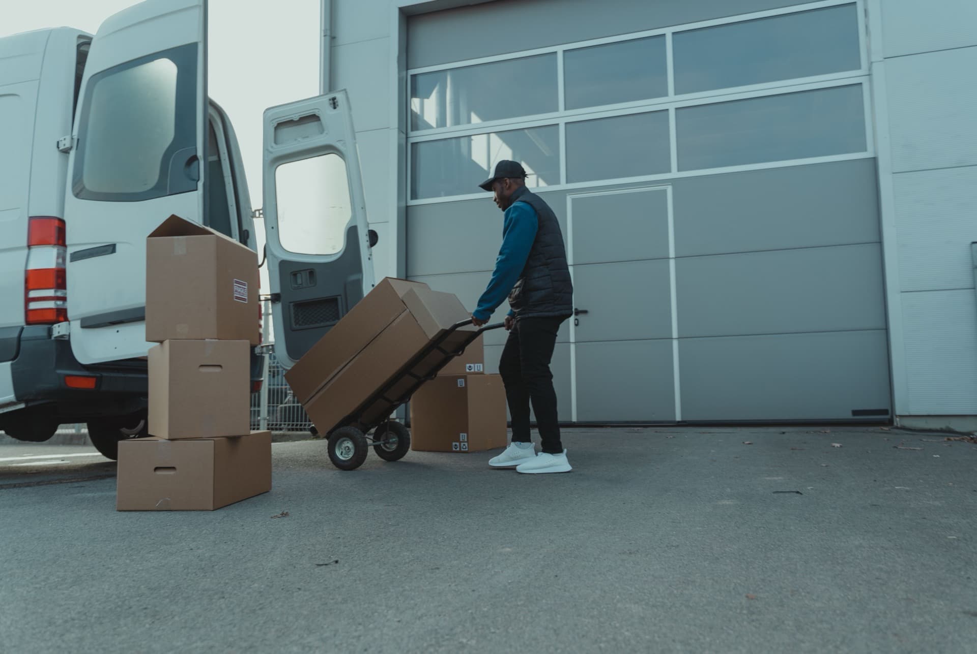 Courier rolling a hand truck of cardboard parcels out of a white cargo van at a warehouse loading dock