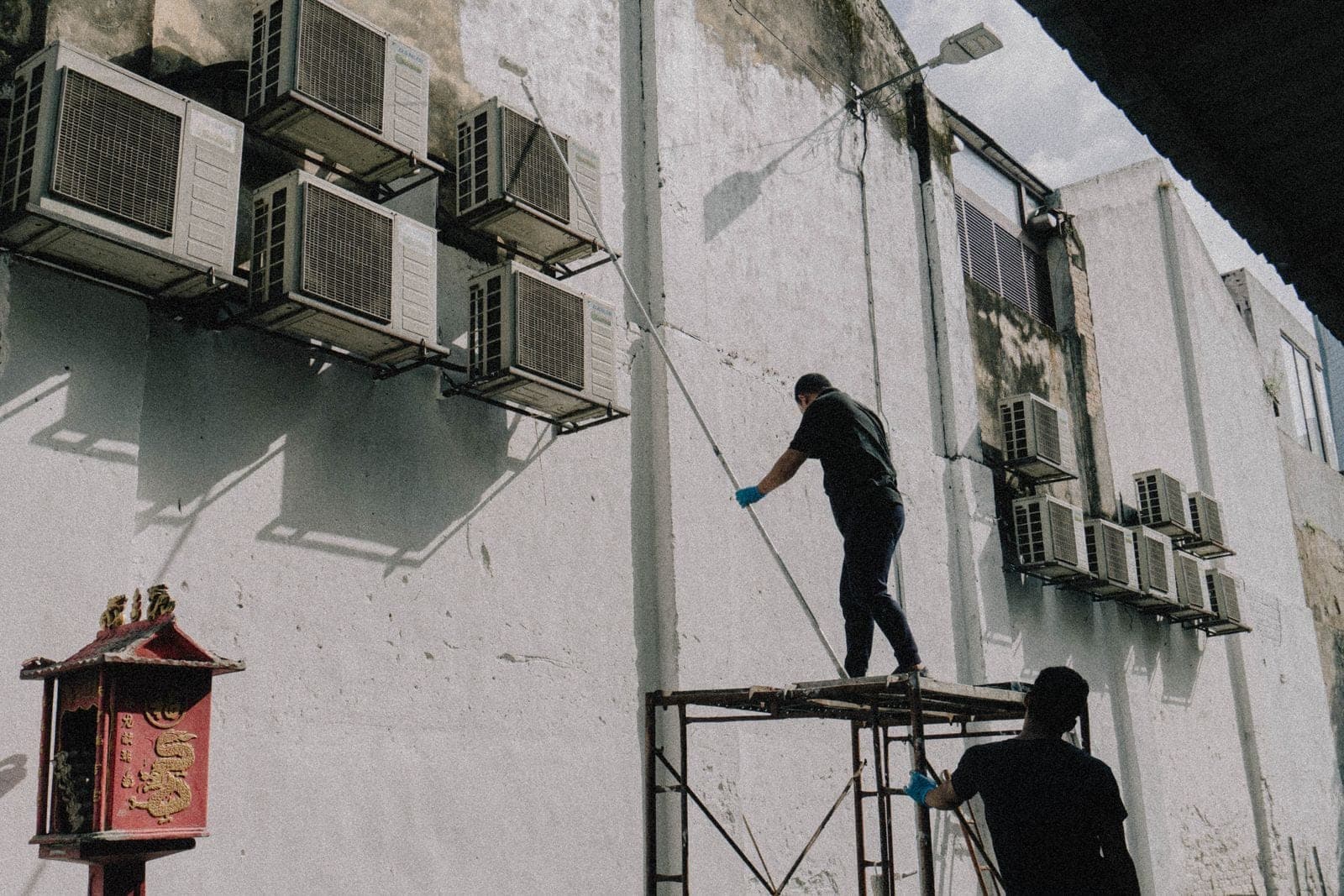HVAC technicians on scaffolding servicing a bank of wall-mounted air-conditioning condenser units on a commercial building