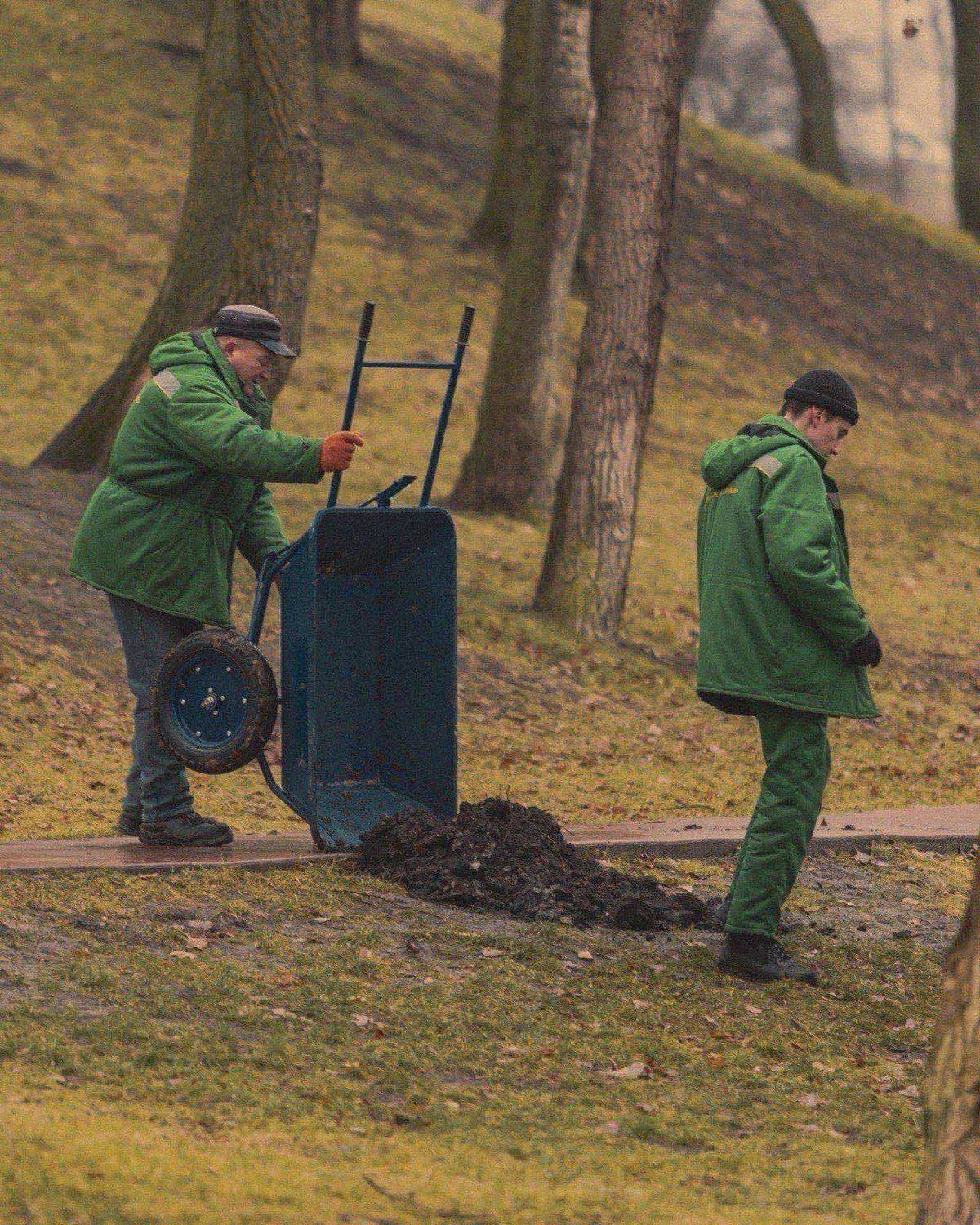 Two landscaping crew members in matching work uniforms spreading mulch from a wheelbarrow in a park setting