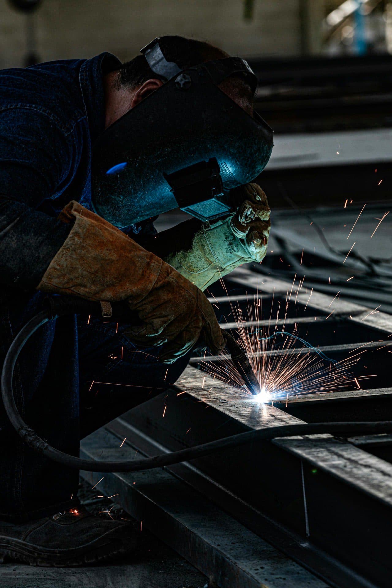 Metal fabricator in a welding helmet joining structural steel with sparks flying on an industrial fabrication floor