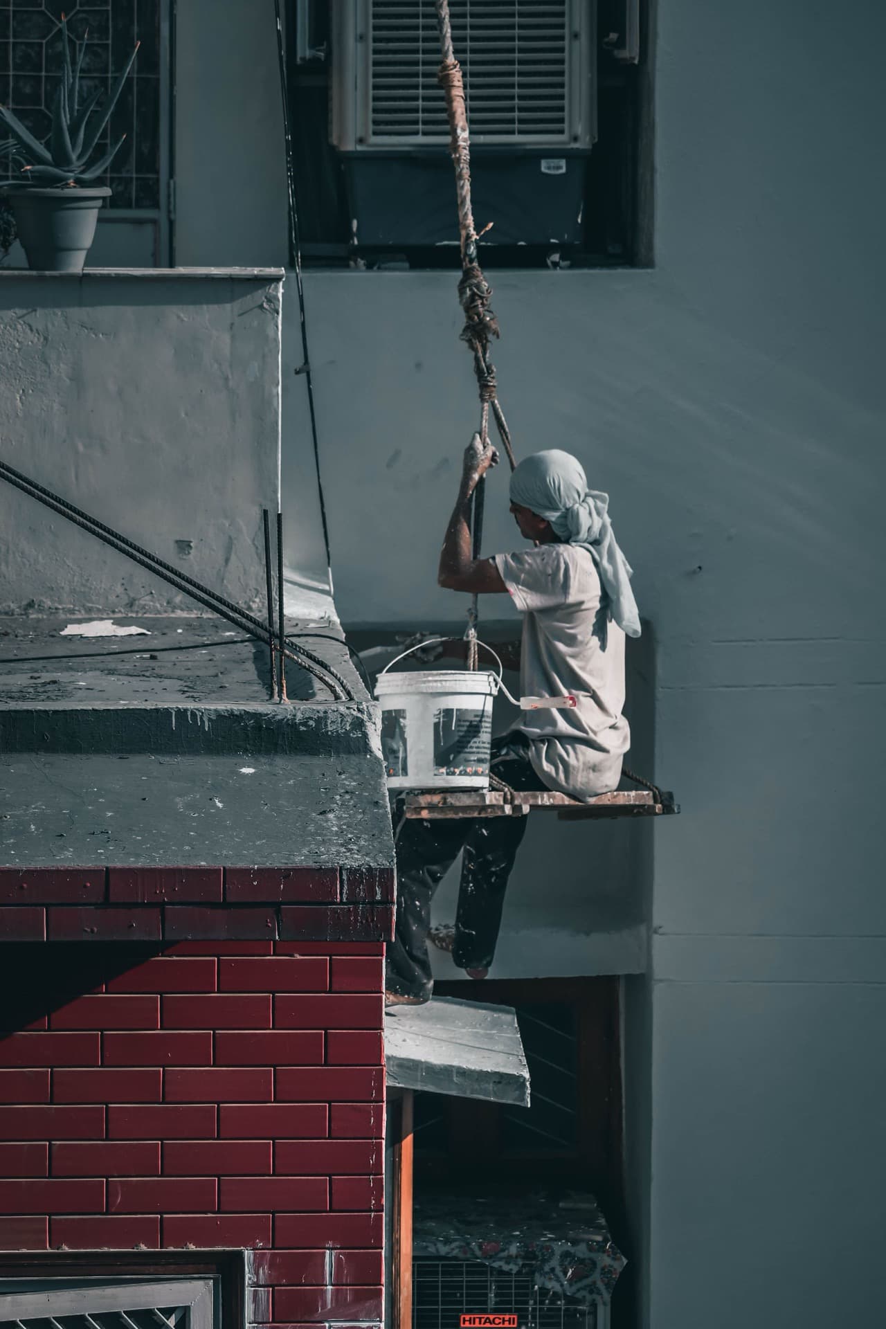 Commercial painter suspended on a rope platform painting the exterior wall of a building, paint bucket at hand