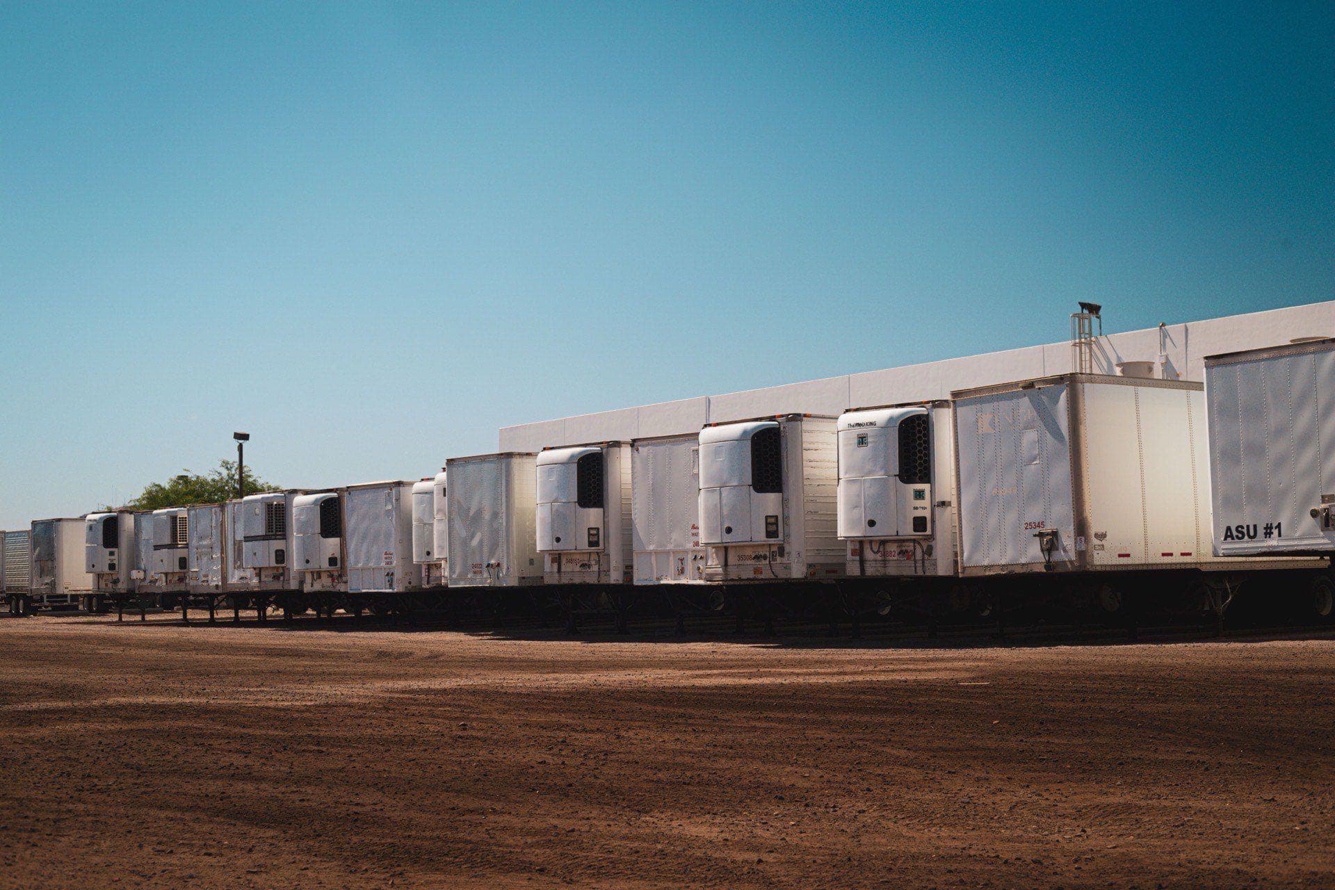Row of white refrigerated semi-trailers with rooftop cooling units parked in a yard under a clear sky